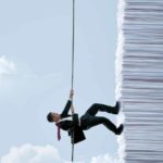 A man in a business suit uses a rope to climb up the side of a huge pile of papers fashioned like a tall building against a blue sky backdrop with clouds representing an assessment of whether CBA shares stacked up well in March