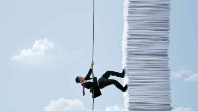A man in a business suit uses a rope to climb up the side of a huge pile of papers fashioned like a tall building against a blue sky backdrop with clouds representing an assessment of whether CBA shares stacked up well in March