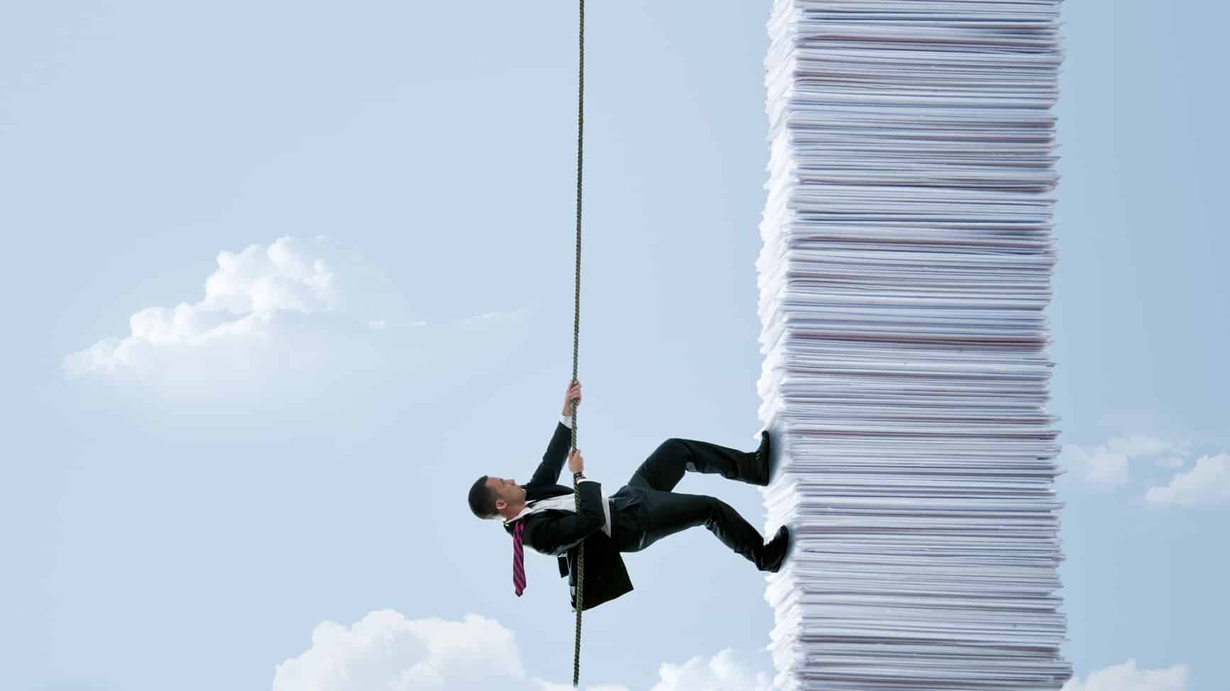 a man in a business suit uses a rope to climb up the side of a huge pile of papers fashioned like a tall building against a blue sky backdrop with clouds.