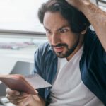 a man sitting in an aeroplane seat holds the top of his head as he looks at his airline ticket with an annoyed, angry expression on his face.