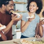Two parents and two children happily eat pizza in their kitchen.