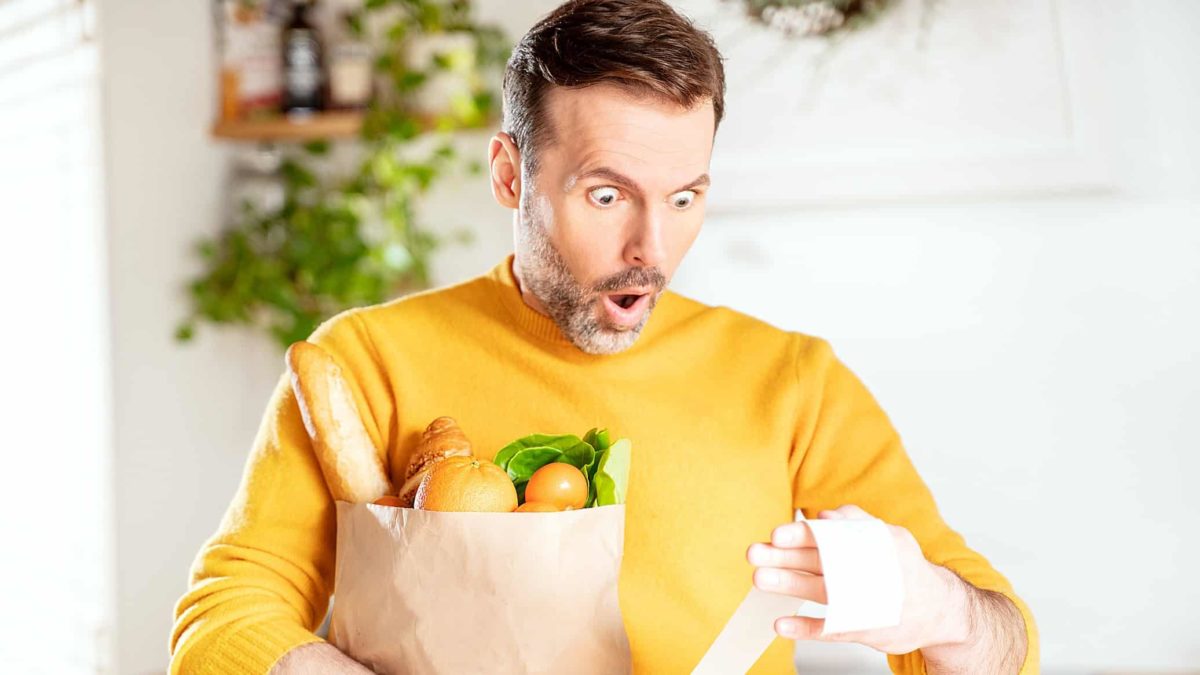 A man holding a paper bag full of food items looks in shocked dismay at his supermarket docket as if high prices have taken him by surprise.