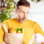 A man holding a paper bag full of food items looks in shocked dismay at his supermarket docket as if high prices have taken him by surprise.