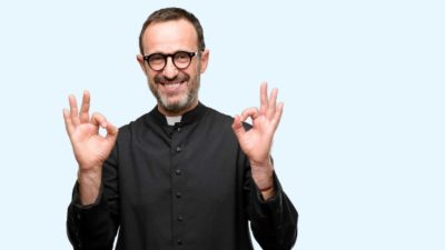 A priest in robes and collar smiles widely and holds up his forefingers and thumbs in circular OK signals.