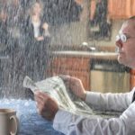 A man sits at his kitchen table reading the paper and drinking coffee as rain pours on him, while a woman stands with an umbrella over her head in the distant background.