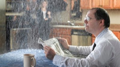 a man sits at his kitchen table reading the paper and drinking coffee as rain pours on him, drenching his shirt and all around him while a woman stands with an umbrella over her head in the distant background, not sharply visible through the rain.