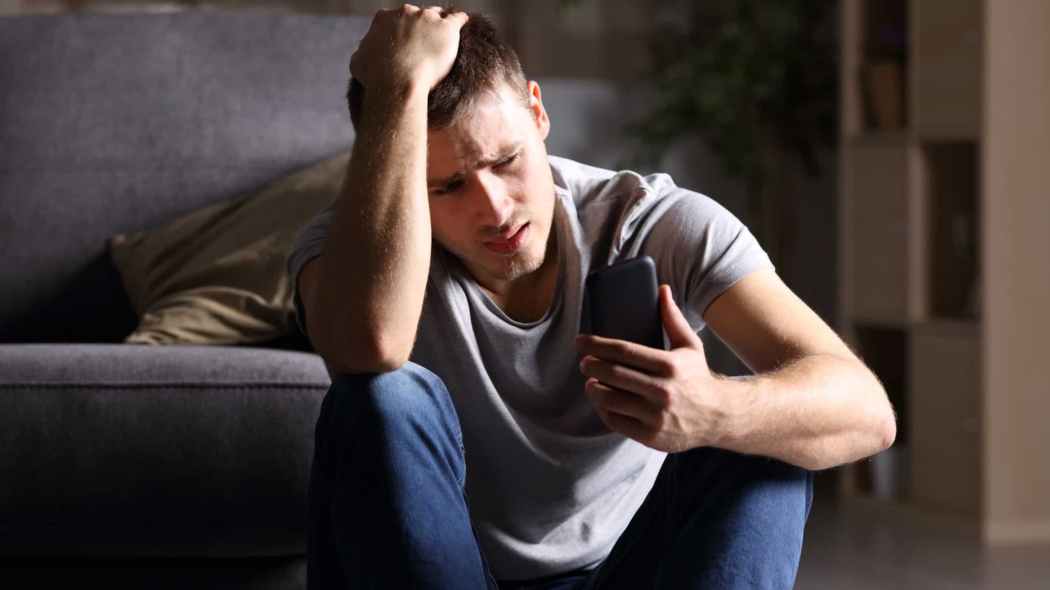 a young man sits on the floor with his back against a sofa hunched over his phone in one hand and his other hand on top of his head as though he is seeing bad news as his face looks sad and anguised.