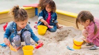 A group of three little girls play together in a sand pit with buckets and spades, each intently concentrating on their own digging projects.