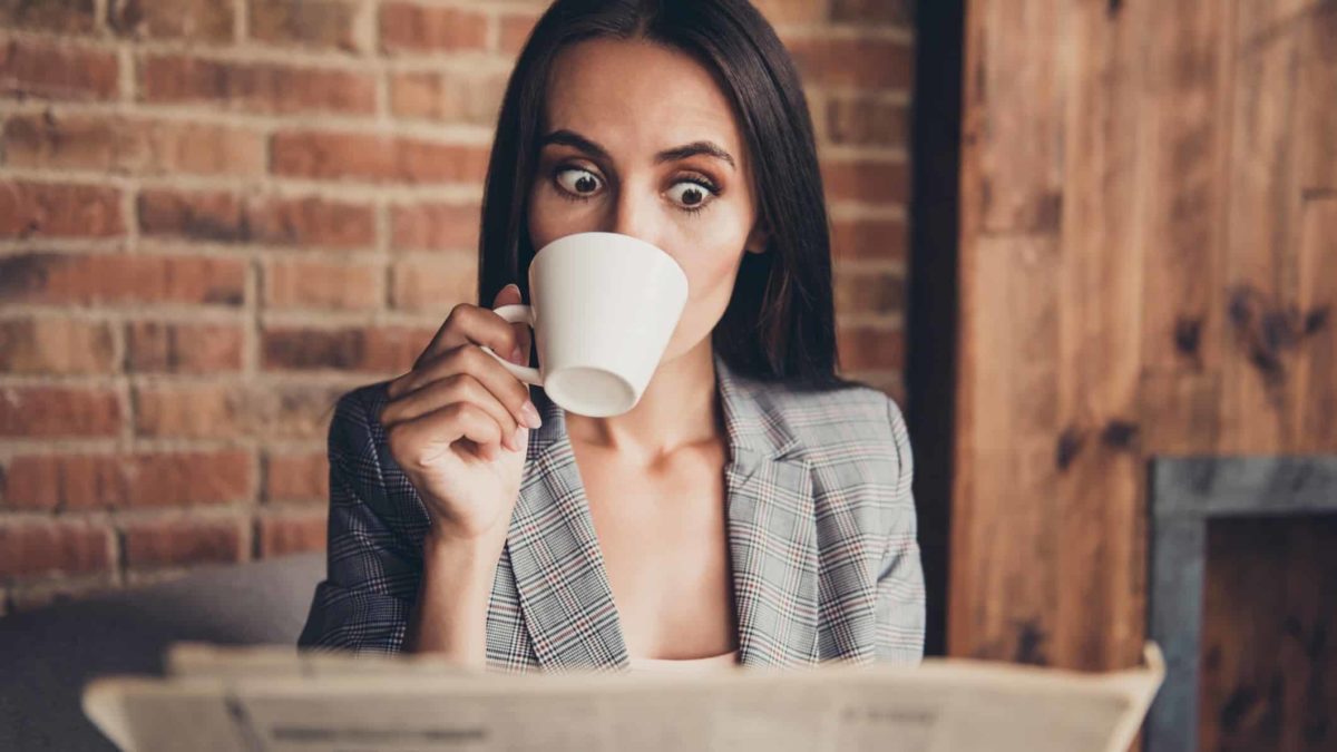A woman looks shocked as she drinks a coffee while reading the paper.