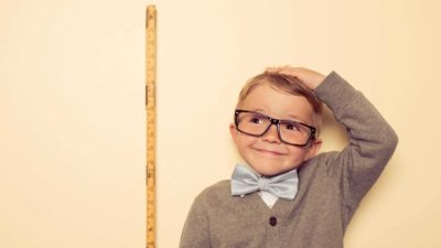 A short boy wearing big glasses stands next to a measuring stick with his hand on his head wondering if he'll ever stop being short, similar to the Polynovo share price which is among the most shorted shares on the ASX right now