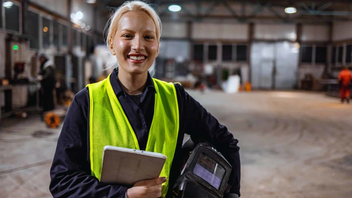 a female steel worker wearing a high visibility vest with her protective helmet tucked under her arm smiles as she carries a clipboard in a large warehouse of steel products.