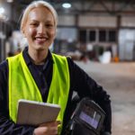a female steel worker wearing a high visibility vest with her protective helmet tucked under her arm smiles as she carries a clipboard in a large warehouse of steel products.