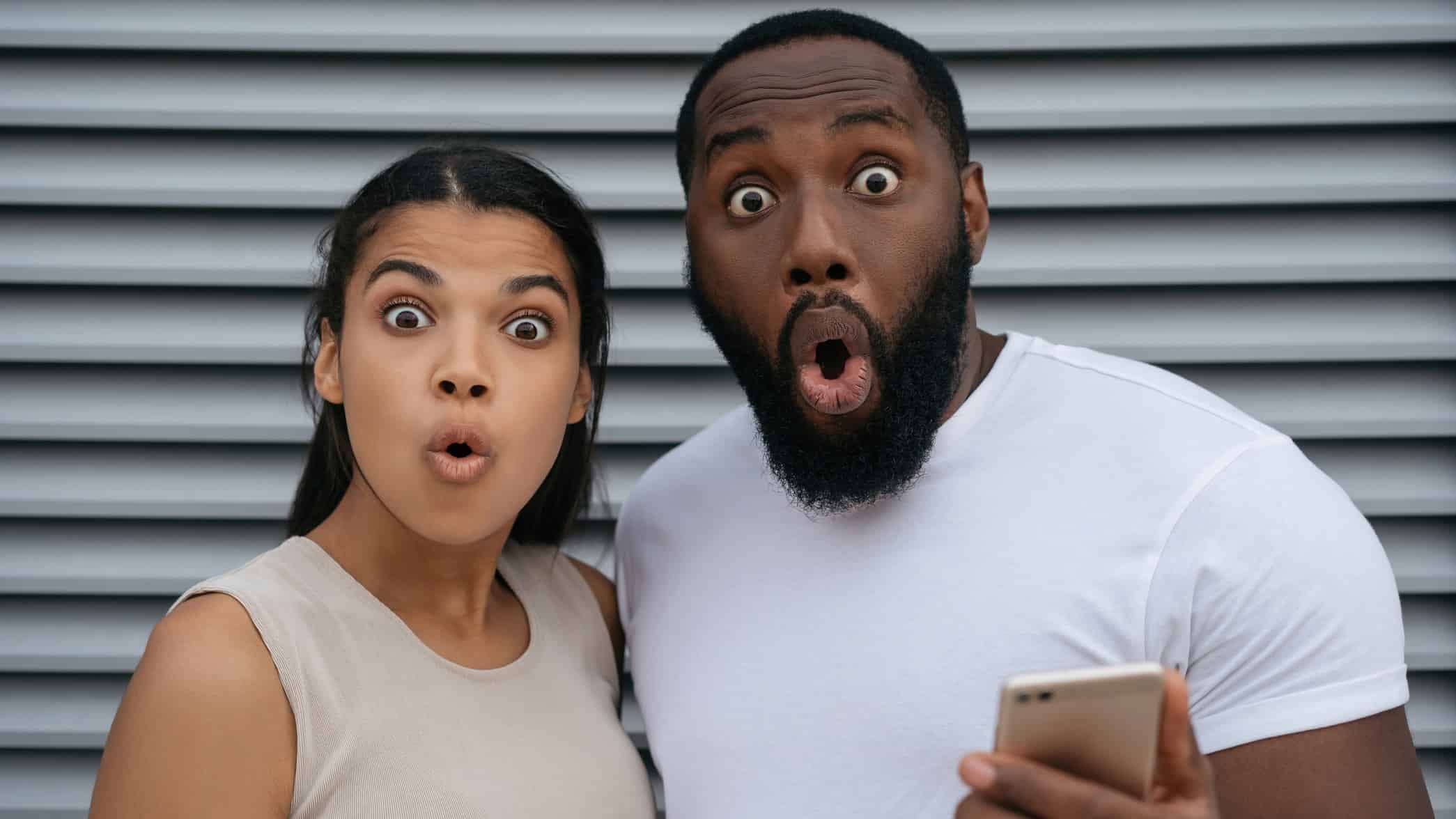 a couple look dumbfounded with exaggerated looks of surpirse on their faces as te mman holds a phone in his hand.