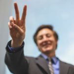 A businessman in soft-focus holds two fingers in the air in the foreground of the shot as he stands smiling in the background against a clear sky.