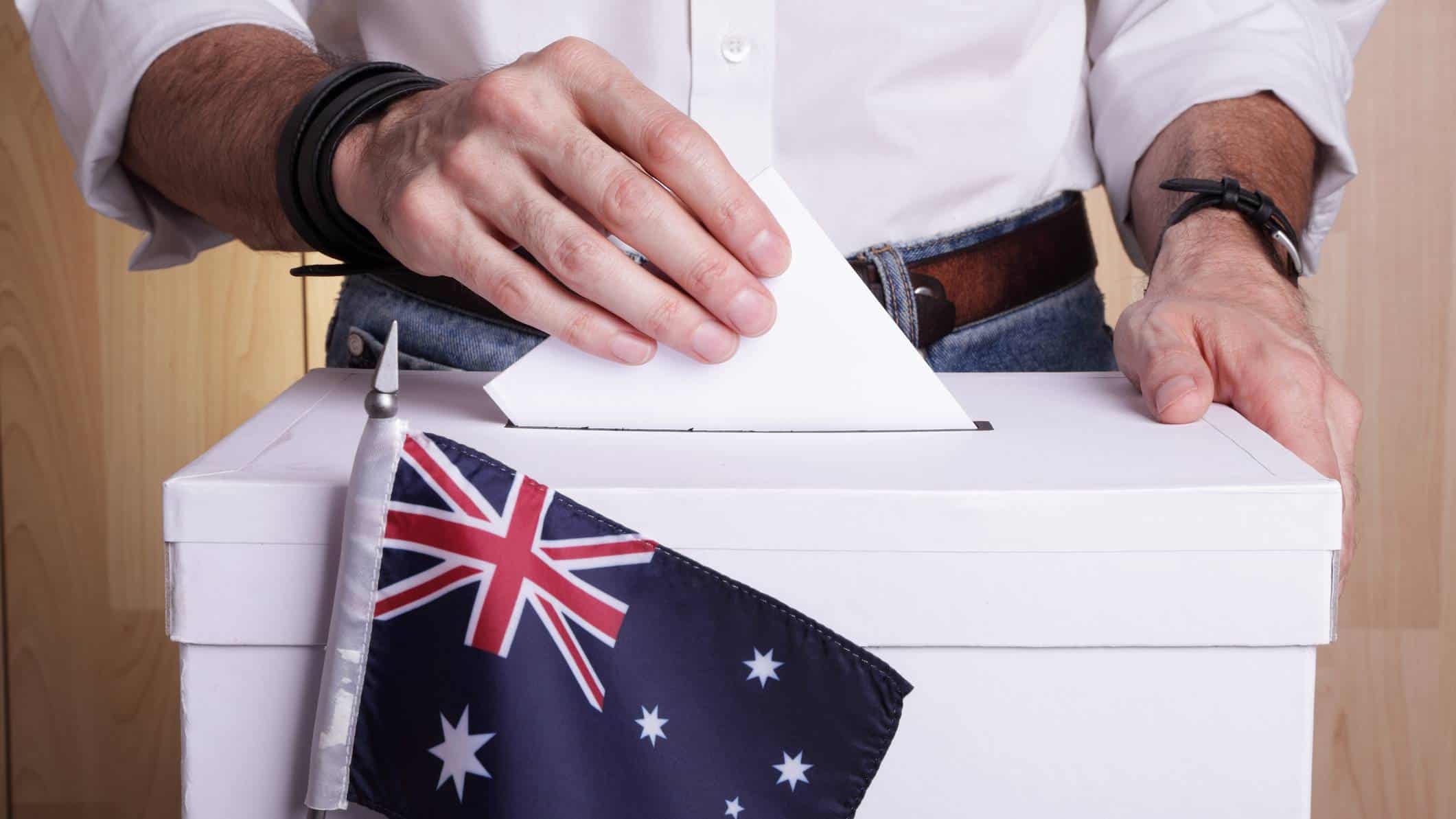 A close-up photo of a ballot box with an Australian flag in front of it and a gentleman's hands placing his vote in the 2022 election inside the box