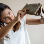 A woman holds her empty unzipped wallet upside down and dips her head to look under it to see if any money falls out of it.