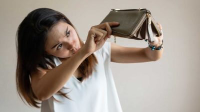 A woman holds her empty unzipped wallet upside down and dips her head to look under it to see if any money falls out of it.