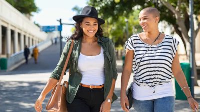 a fashionable older woman walks side by side with a stylish younger woman in a street setting as they both smile at something they are talking about.