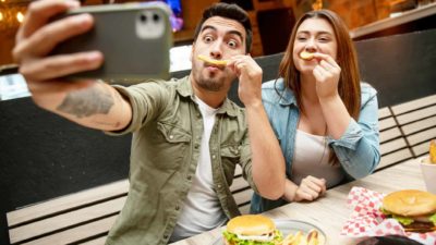 A couple makes silly chip moustache faces and take a selfie on their phone.