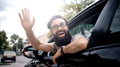 A man leans out of his car window with a massive smile on his face and waves.