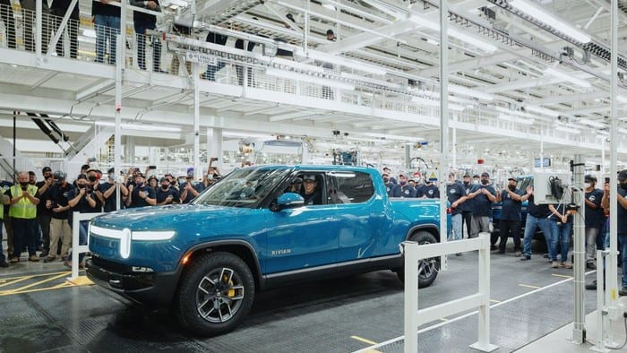 A blue utility truck takes pride of place in an auto factory setting with hundreds of workers gathered around admiring it.