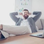 A man leans back with his hands behind his head and feet on his desk with a big smile on his face at his success.
