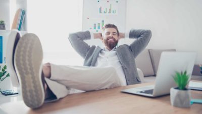 A man leans back with his hands behind his head and feet on his desk with a big smile on his face at his success.