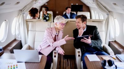 Two people in first class of an aeroplane share advice over the aisle of the plane.