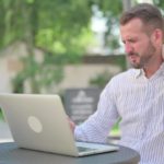 A man sits uncomfortably at his laptop computer in an outdoor location at a table with trees in the background as he clutches the back of his neck with a wincing look on his face.