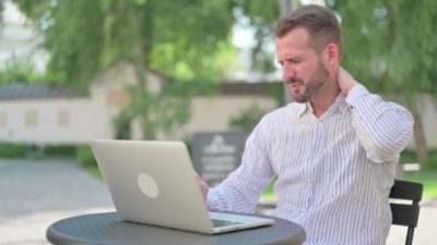 A man sits uncomfortably at his laptop computer in an outdoor location at a table with trees in the background as he clutches the back of his neck with a wincing look on his face.