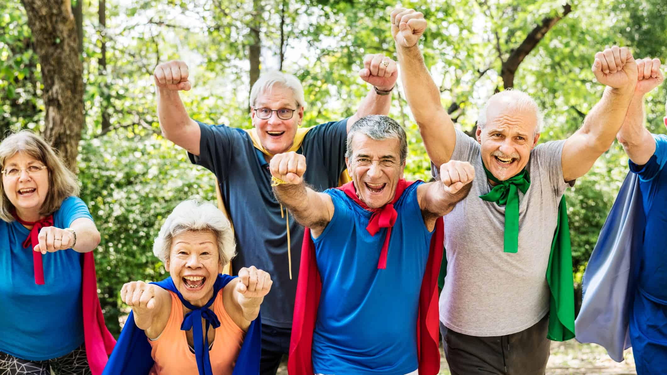 A group of older people wearing super hero capes hold their fists in the air, about to take off.