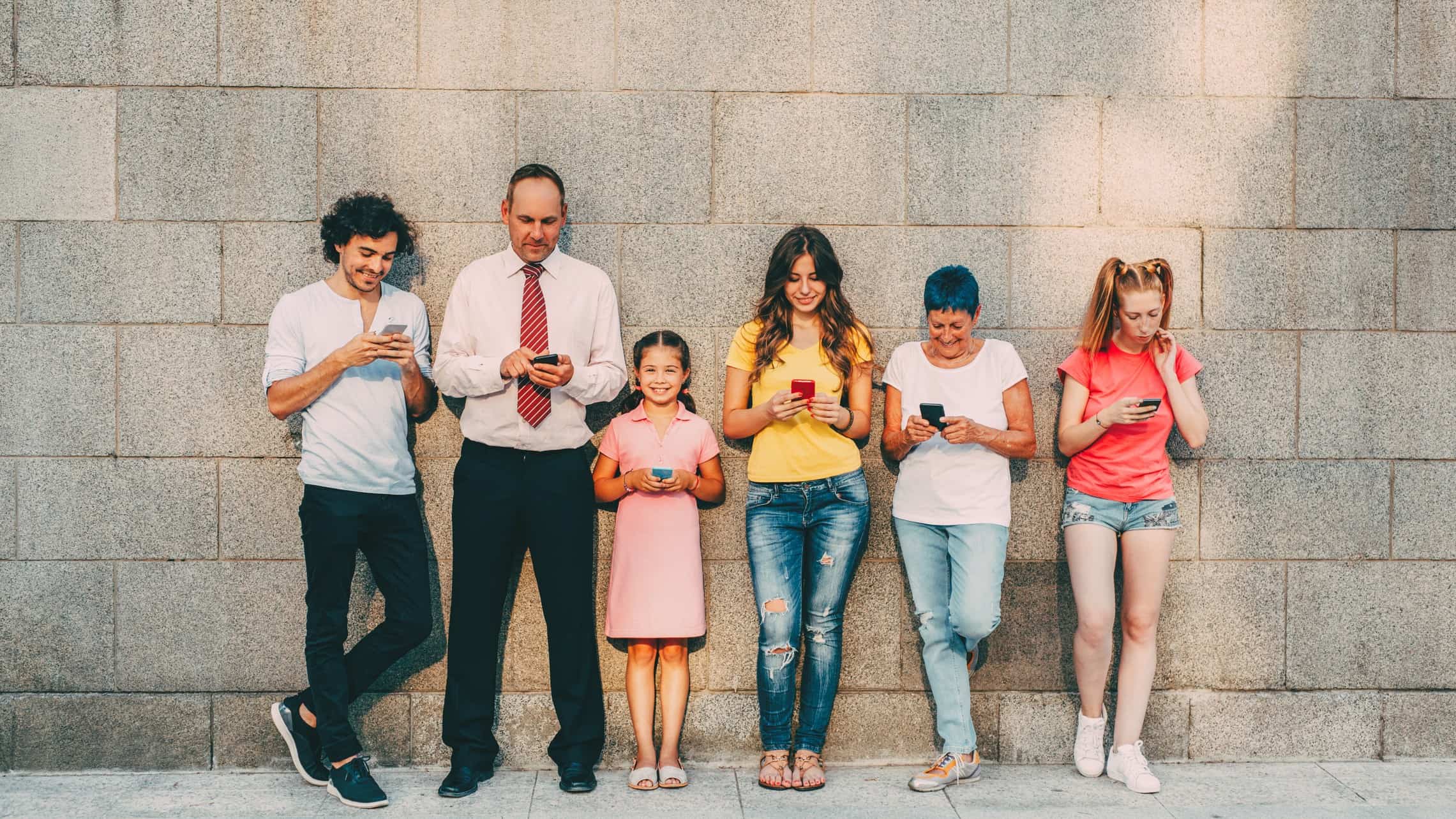 A group of people of all ages, size and colour line up against a brick wall using their devices.