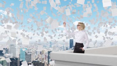 A woman stands on the roof of a city building as papers fly in the sky around her.