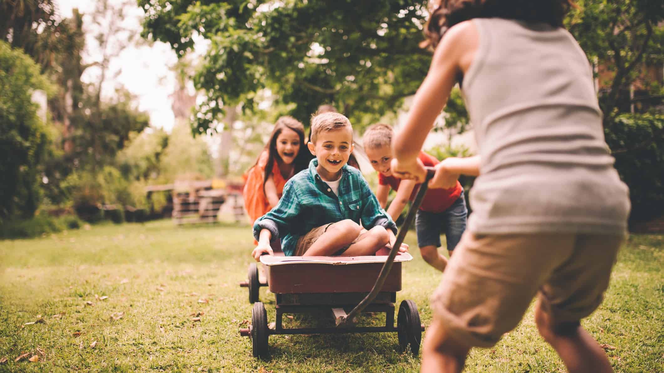 A kid pulls his friends on a wagon in the backyard.