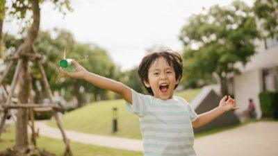 A little boy runs around the playground lifting a toy aeroplane in the air above his head.