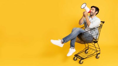 A man sits in a shopping trolley and shouts buy through a megaphone.