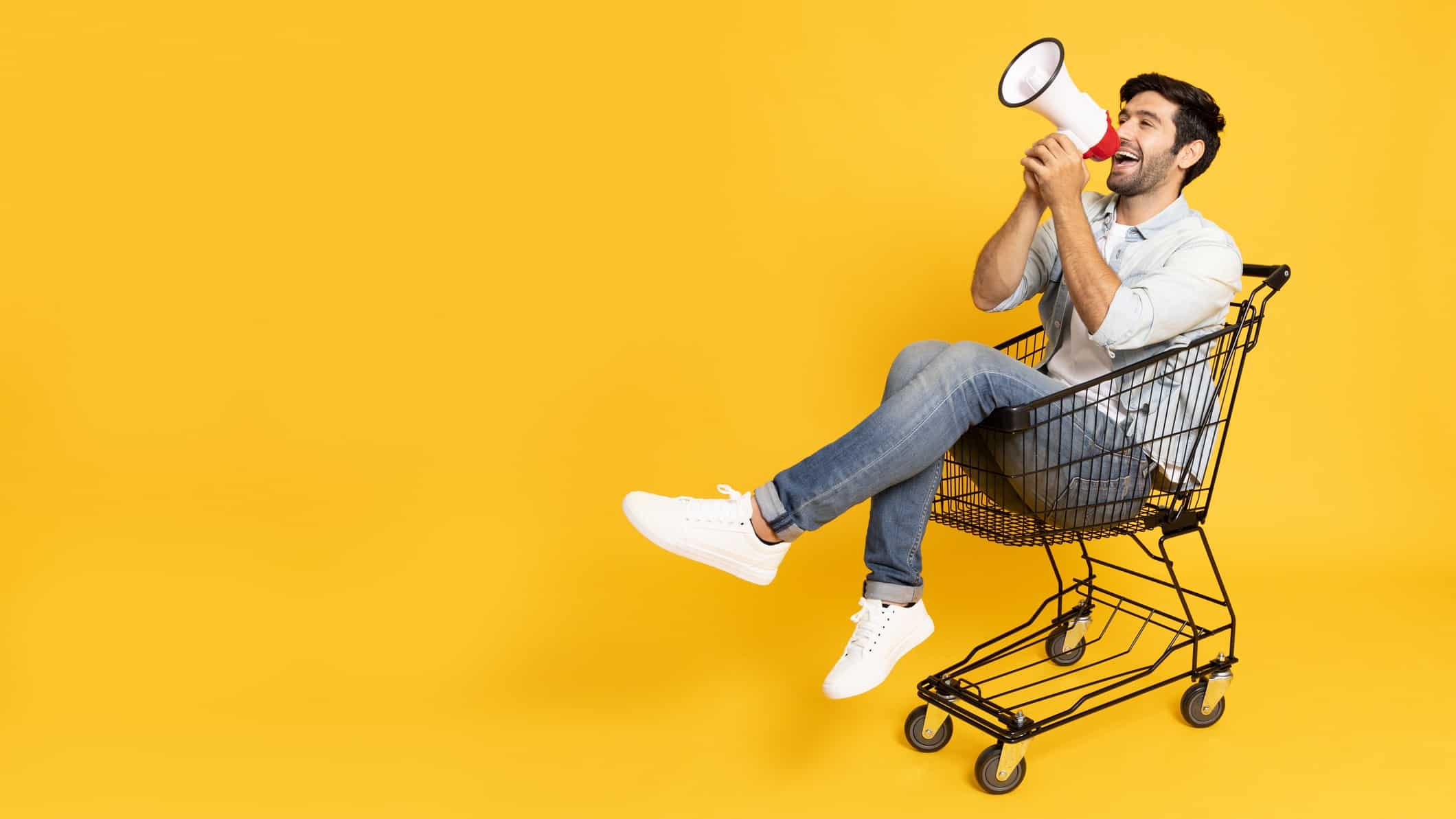 A man sits in a shopping trolley and shouts buy through a megaphone.