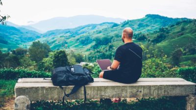 A man sits on a bench atop a mountain with a laptop, making investments with a green ESG mind.