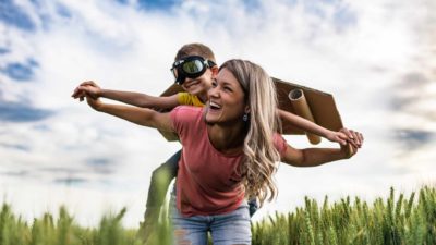 A little boy in flying goggles and wings rides high on his mum's back with blue skies above.