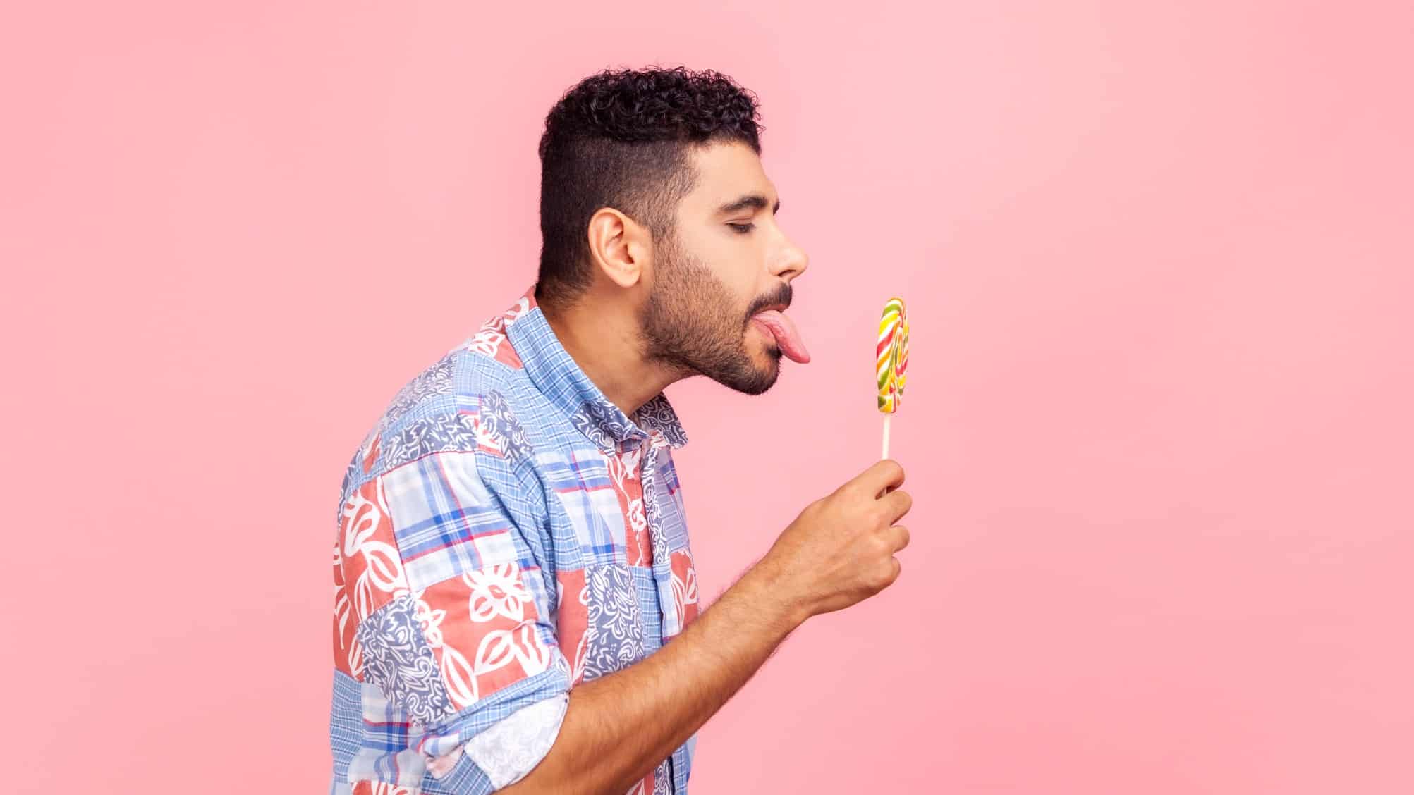 A man in a colourful party shirt sticks his tongue out really far to lick a lollipop.