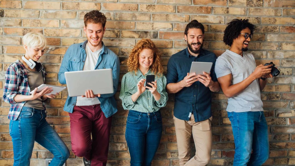 A group of young people lined up on a wall are happy looking at their laptops and devices as they invest in the latest trendy stock.