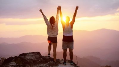 Two people climb to the summit and raise their arms in success as the sun rises brightly over the mountains.