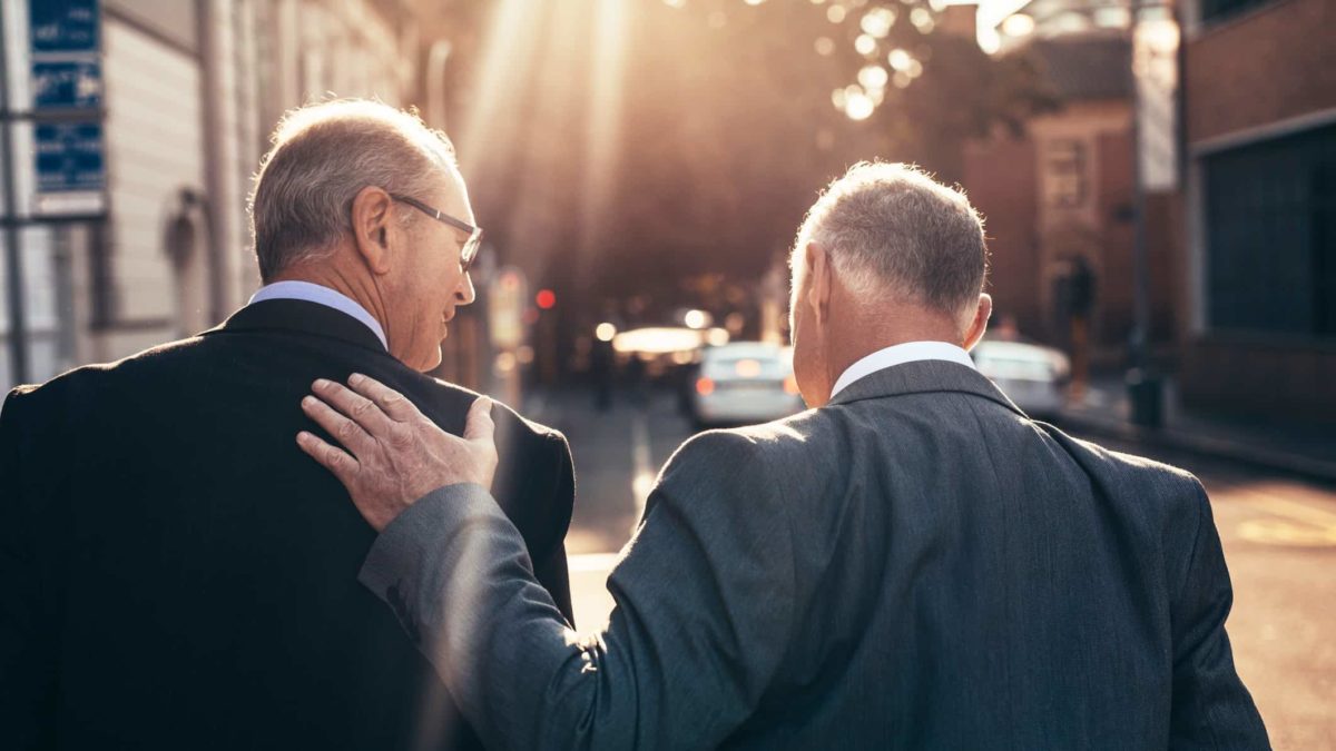 Two older men in suits walk down the street in the sunlight, one congenially rests his hand on the other's shoulder.
