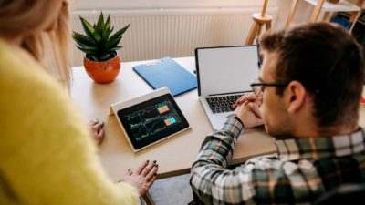 A man and woman watch their device screens, making investing decisions at home.