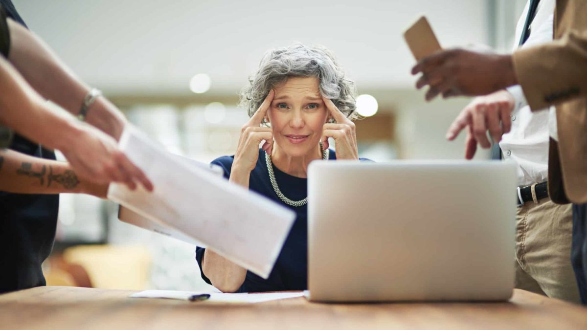A mature businesswoman has people demanding things from her from all angles, and she looks stressed.