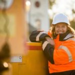 A woman working in construction leans against a piece of machinery wearing a hi vis vest and a hardhat, smiling.