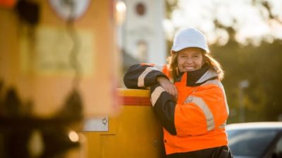 A woman working in construction leans against a piece of machinery wearing a hi vis vest and a hardhat, smiling.