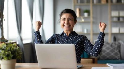 A woman gives two fist pumps with a big smile as she learns of her windfall, sitting at her desk.