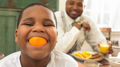 A young boy smiles with a juicy slice of orange in his mouth, eating breakfast at the dining table with his dad.