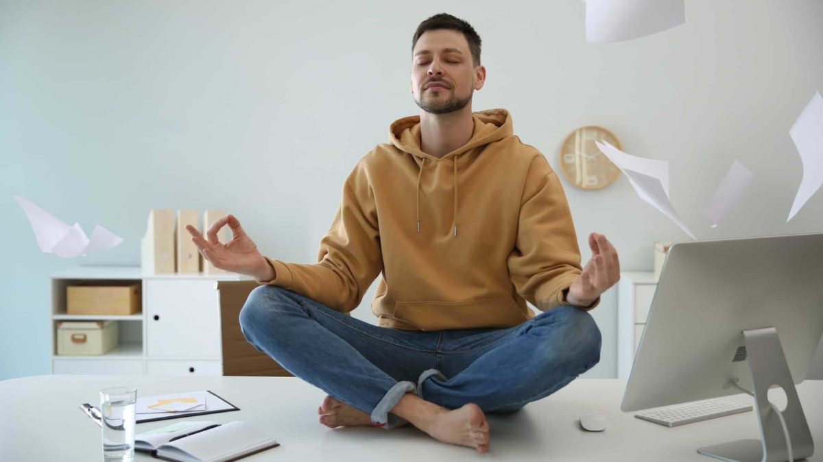 A man sits cross-legged in a zen pose on top of his desk as papers fly around his head, keeping calm amid the volatility.